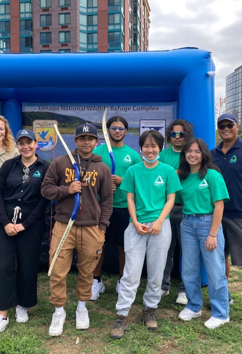 8 people stand in front of a large inflated square object. Some of the people hold archery bows. The group is smiling. It's a cloudy day and there are urban buildings in the background.