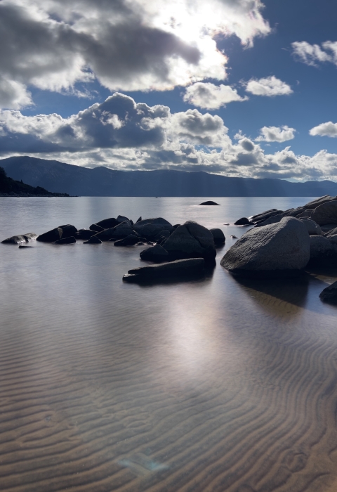 A lake with boulders in the forefront and mountains in the distance as clouds pass over the sun