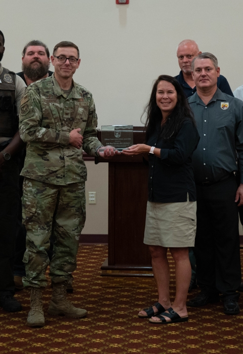 A man in fatigues accepts an award from a woman in a U.S. Fish and Wildlife Service shirt with a group of other people standing around them