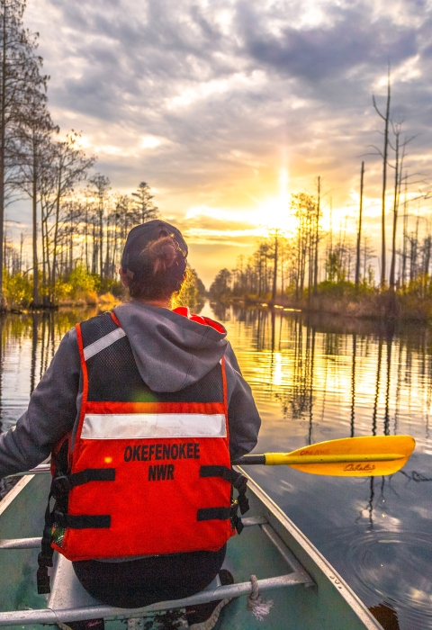 woman in front of canoe holding a yellow paddle in dark reflective water with cypress trees and clouds in a sunny sky. 