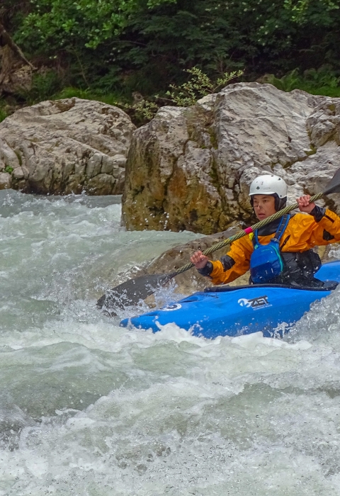 A woman in a blue kayak paddles down white water. She wears a white helmet, orange jacket, and blue bib.