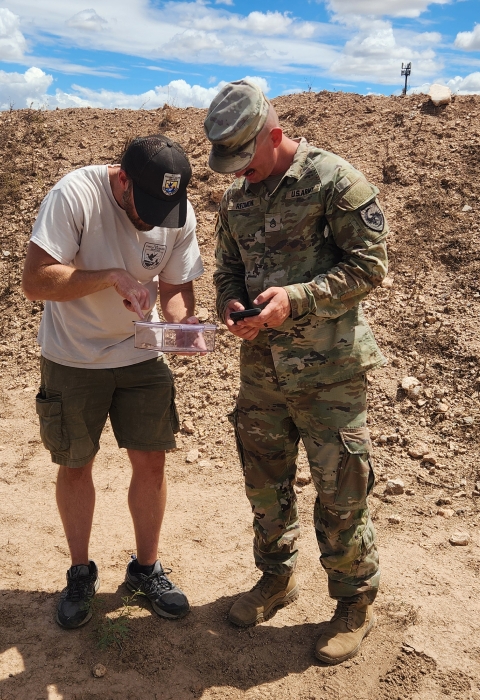 a fish and wildlife service biologist and military personnel dressed in fatigues looks down at a container with a lizard