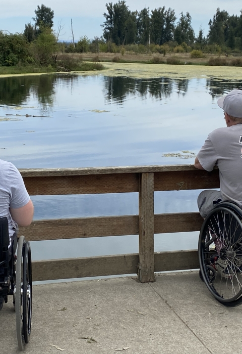 Two men in manual wheelchairs sit side by side fishing from a paved path, looking out over a calm pond bordered by trees and greenery, with a wooden railing in front of them.