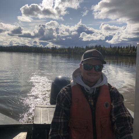 A person wearing an orange lifejacket, flannel shirt, and hooded sweater, along with a hat and sunglasses,smiles as they steer a boat's rear engine on a river under a partly cloudy sky.