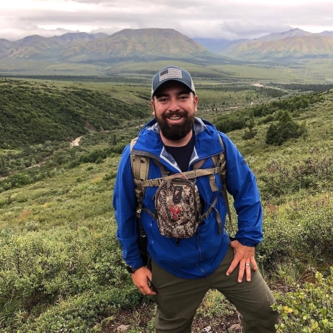 A person stands on a side of a hill posing for a picture with a binocular chest harness on and a hat with a rain coat on. Mountains on the horizon and a valley in between are visible. 