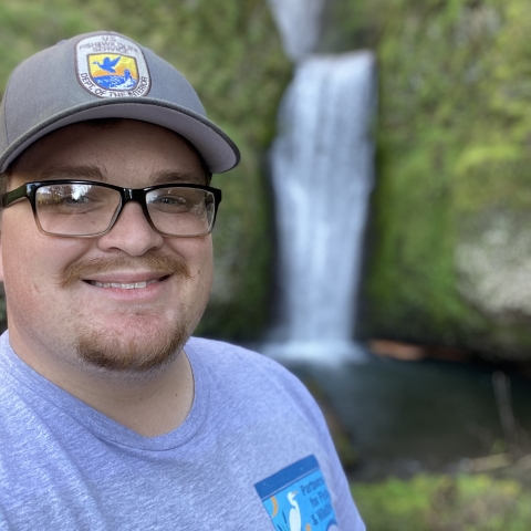 A person wearing glasses and a hat looks directly at the camera. The shirt features the Partners for Fish and Wildlife Program signature on the upper left, and the hat displays the U.S. Fish and Wildlife Service emblem. Behind the person, an out-of-focus waterfall cascades down a rocky surface.
