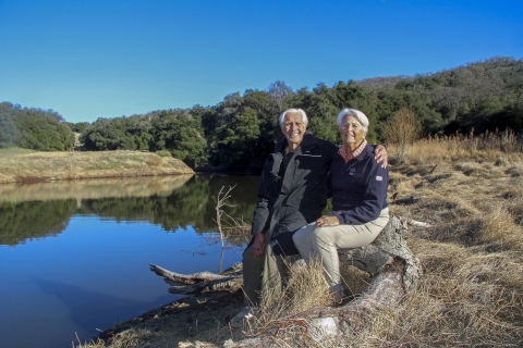 Two elderly people sitting on a log next to a body of water pose for a picture
