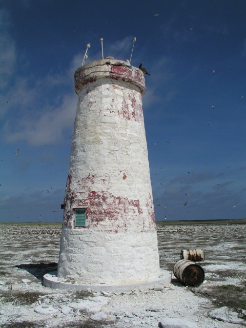 The ruins of a lighthouse. It is white with faded splotches of red. Around it sits two metal drums. 