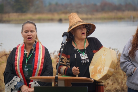 Two native singers, with one wearing a straw hat and holding a drum, behind a podium