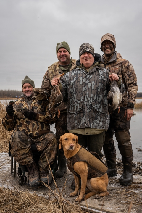 four adult hunters with wetland in background pose in camouflage hunting gear with dog