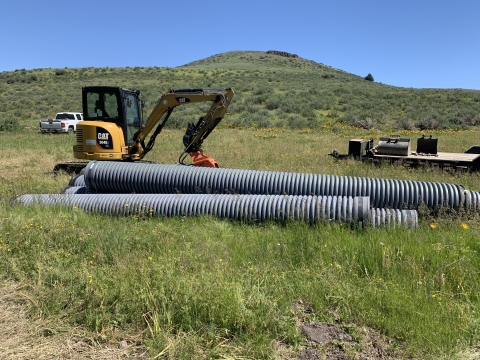 4 large gray pipes are seen in the grass in front of a backhoe machine.