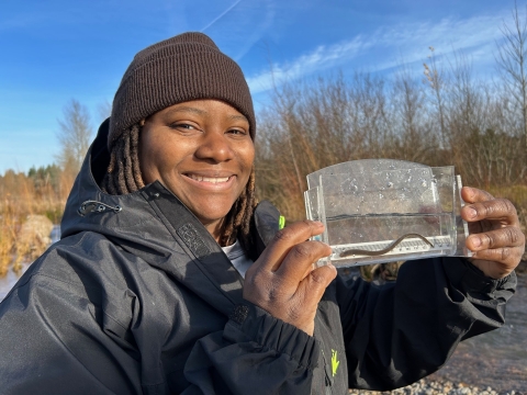 A woman wearing a beanie and jacket holds up a clear container with water and a small aquatic organism