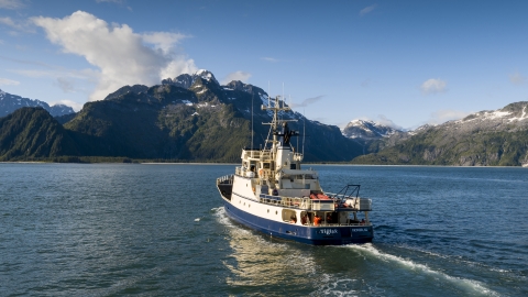 Blue and white ship sails towards land over calm blue waters and a blue sky with a few clouds.
