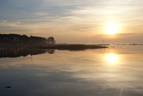 The sun goes down on a snowless but wintry-looking day and reflects against the blue-gray water of the Blackwater River in the image's foreground. On the horizon line, a stand of trees and marsh grass is visible. 