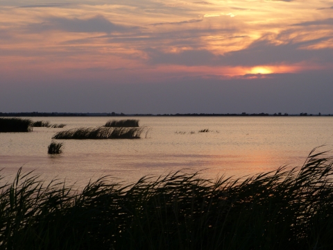 A setting sun ducks behind clouds over an expanse of open water dotted with marsh grasses