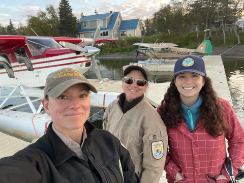 Three people standing on a dock with two single engine float planes at the dock.