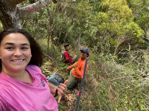 A woman on a steep wooded slope poses for a selfie. Two people smile in the background.