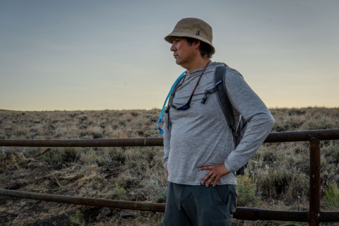 Man stands in front of a fence line with sagebrush in the background.