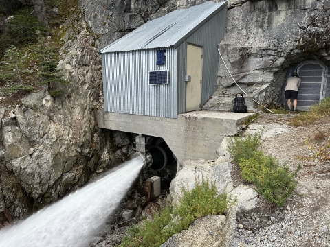 A large quantity of water shoots out of a mountain trhough a pipe. A shed sits over it, and a person opens a door to a tunnel next to it.