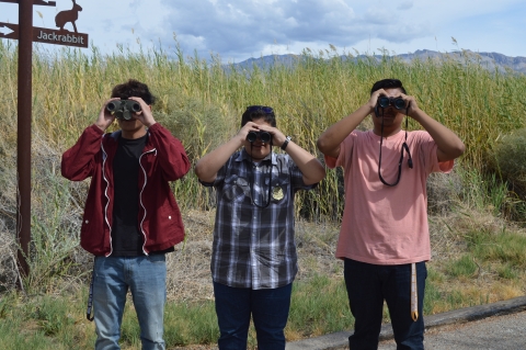 Three students looking through binoculars posing in front of a grassy field