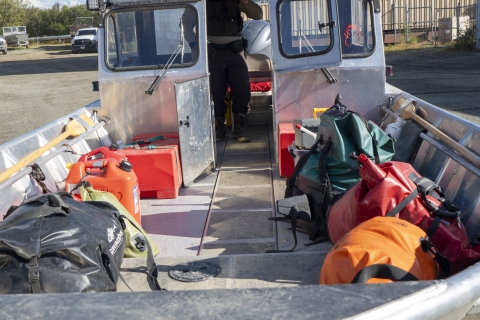 a view onto a riverboat deck loaded with dry bags filled with gear and extra fuel containers. The boat is on a trailer in a parking lot. A uniformed officer stands inside the enclosed cabin at the stern of the river boat. 
