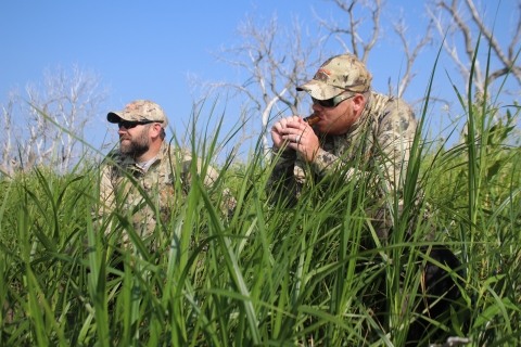 Two men dressed in camouflage sitting in grass. One man is holding a duck call to has mouth.