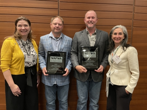 Four people are standing in the frame. From left to right, there is a woman with a yellow sweater and brown hair, then a man in a blue blazer holding a plaque award, next to another man holding an award, and the last person is a woman in a white blazer with salt and pepper colored hair. 