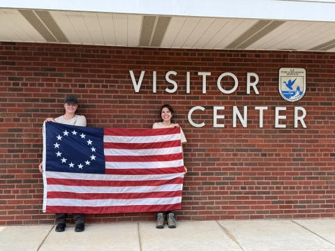 2 people hold up Betsy Ross flag in front of brick wall saying Visitor Center