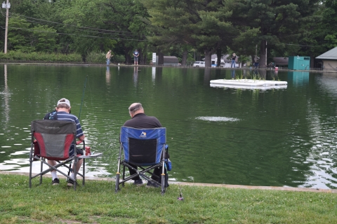 Two men seated in chairs on the shore of a pond fishing, with a floating wetland in the distance