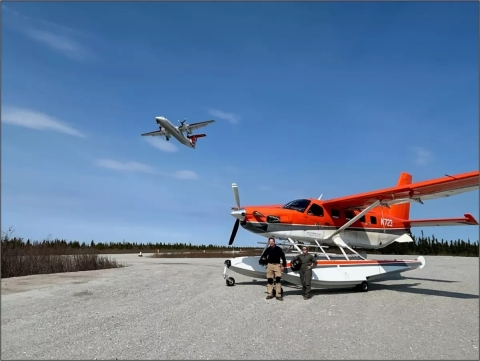 plane flying over two pilot biologists standing in front of a plane
