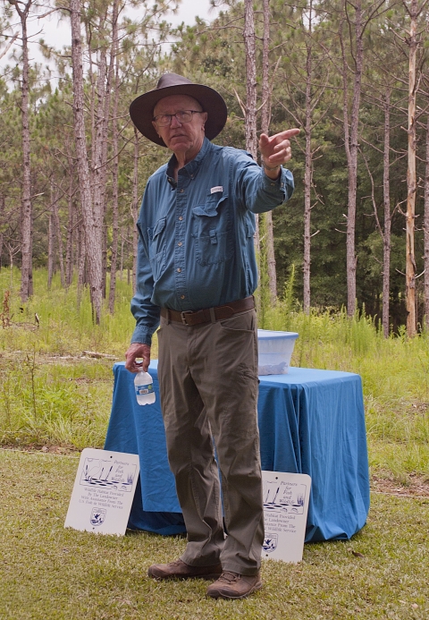 A man gestures to the left while standing beside a table covered with a blue tablecloth, set in front of longleaf pine trees and a grassy understory. At his feet, two signs lean against the table, both reading “Partners for Fish and Wildlife” at the top and “Wildlife Habitat Provided By The Landowner With Assistance From the U.S. Fish and Wildlife Service” beneath.