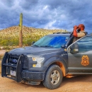 A law enforcement officer standing in his truck in a desert setting looking through binoculars