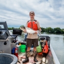 A biologist wearing rubber boots and a brightly colored life vest stands among a pile of carp on a boat while holding a carp.