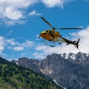 A small yellow helicopter with a crew member looking out soars above rocky mountains in Wyoming.