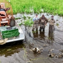 three people stand next to an airboat and a trap full of ducks