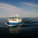 The Research Vessel Connecticut, a 90' steel blue and white ship with a tall mast and various antennas, is traveling through the water with no land in sight. The horizon stretches out far in the distance. The sky is blue, with white clouds.