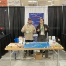 Two biologist stand smiling behind an outreach booth focused on invasive species prevention and identification in Alaska.