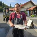 Addison Emerson holding an adult coho salmon at Quilcene National Fish Hatchery. Forklift, facility, and skyline in the background.