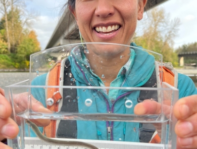 A woman smiles while holding up a clear container holding water and a small, thin eel like fish. The ruler inside measures it around 12 cm in length