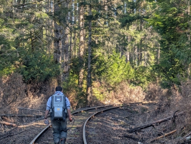 A person wearing a technical backpack walks down railroad tracks in the forest
