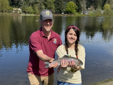 Service staff member and youth event participant posing with a rainbow trout. Lake, houses and trees in the background.