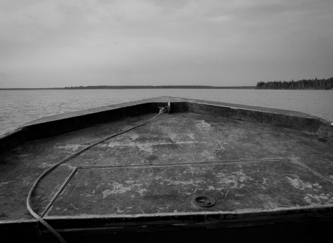 A black and white photo of the nose of a motor boat, taken from within the boat, as it rumbles through the Yukon River. Beyond the boat, and everywhere around it, is water.
