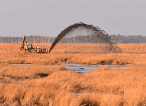 A machine operator sprays a black arc of sediment into the marsh.