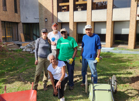 Five people stand in the yard of a building with wheelbarrows and other gardening tools