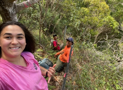 A woman on a steep wooded slope poses for a selfie. Two people smile in the background. 