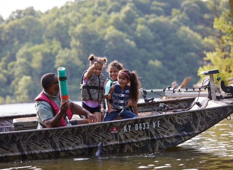 A man is fishing in a boat with three young girls. The kids are excitedly pulling a fish out of the water. 