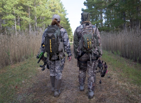 Two hunters in camouflage holding compound bows walk away from the camera down a wide path through a pine forest.