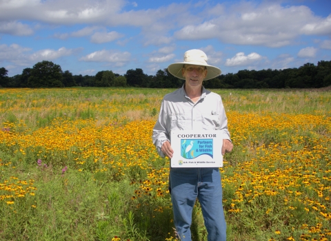 A person wearing a wide‑brimmed hat stands in a large field filled with yellow wildflowers. They hold a sign that reads “Cooperator – Partners for Fish & Wildlife – U.S. Fish & Wildlife Service.” Trees and a blue sky with scattered clouds appear in the background.